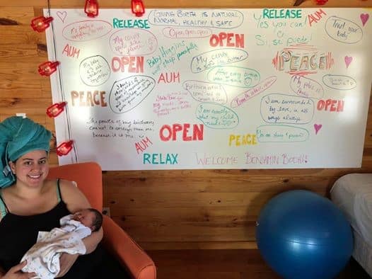 Hypno-mom sitting in a chair holding her newborn next to a big sign of birth affirmations and her hair up in a towel.